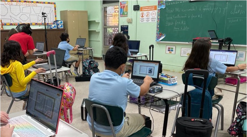 La foto muestra a un grupo de seis estudiantes de escuela elemental sentados en sus pupitres mientras trabajan individualmente en sus computadoras portátiles con la redacción de sus escritos en el salón de clases. La maestra está de pie al lado de una de ellas observando la pantalla.