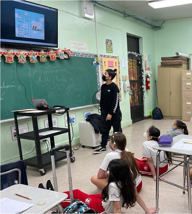 Un estudiante presenta su cuento en la pantalla electrónica del salón de clases, mientras le observan tres compañeras sentadas en el suelo.